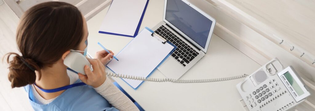Pielęgniarka w maseczce rozmawia przez telefon przy biurku i notuje na klip¬bordzie obok otwartego laptopa